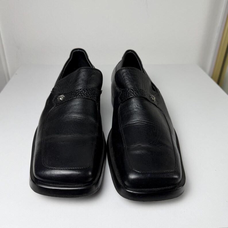 May include: A pair of black leather slip-on shoes with square toes. Each shoe has a strap across the top with a decorative silver emblem. The shoes are displayed on a white surface.
