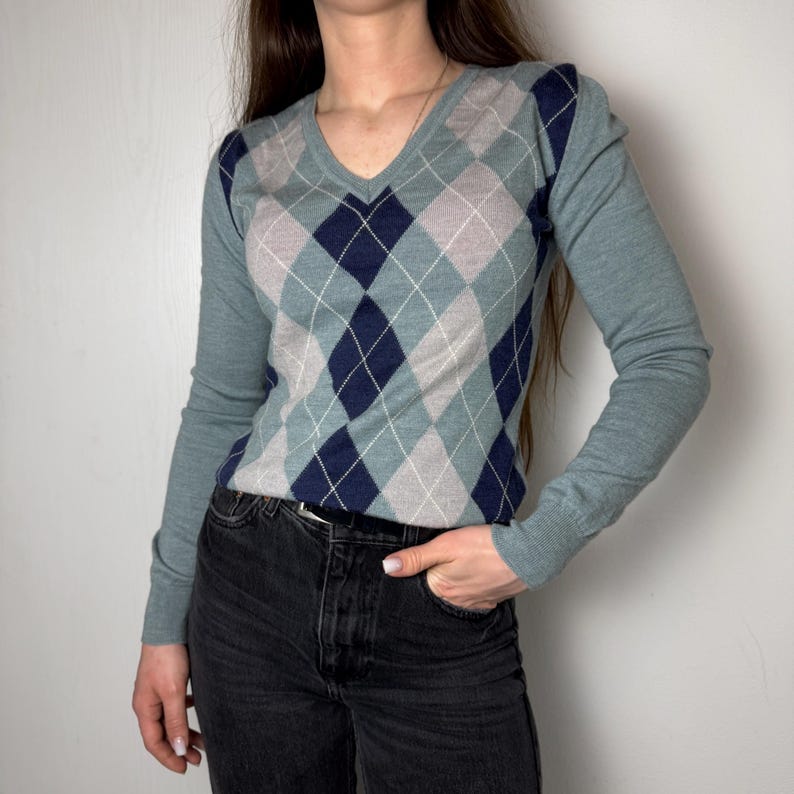 May include: A teal V-neck sweater with a diamond argyle pattern in navy blue and grey. The sleeves and shoulders are plain teal. The sweater is worn with black denim jeans. The argyle pattern is a traditional design.