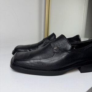 May include: A pair of black leather loafers with a square toe design. The shoes have a textured vamp and a decorative strap with a small emblem. The soles and heels are also black.