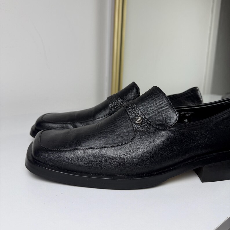 May include: A pair of black leather loafers with a square toe design. The shoes have a textured vamp and a decorative strap with a small emblem. The soles and heels are also black.