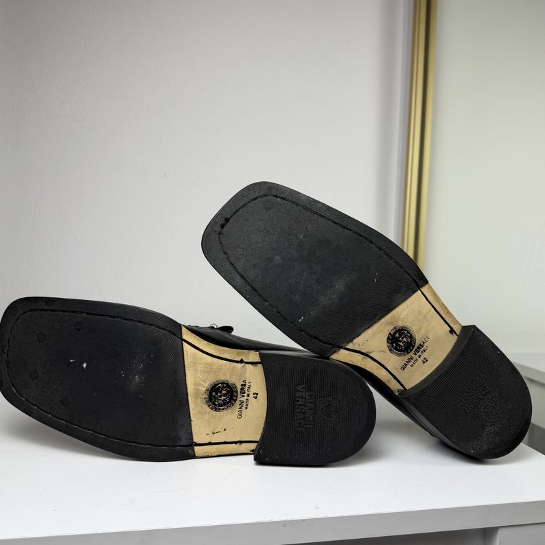 May include: Black leather dress shoes with square toes. The soles are black with a beige heel and the Versace logo. The shoes are size 42.