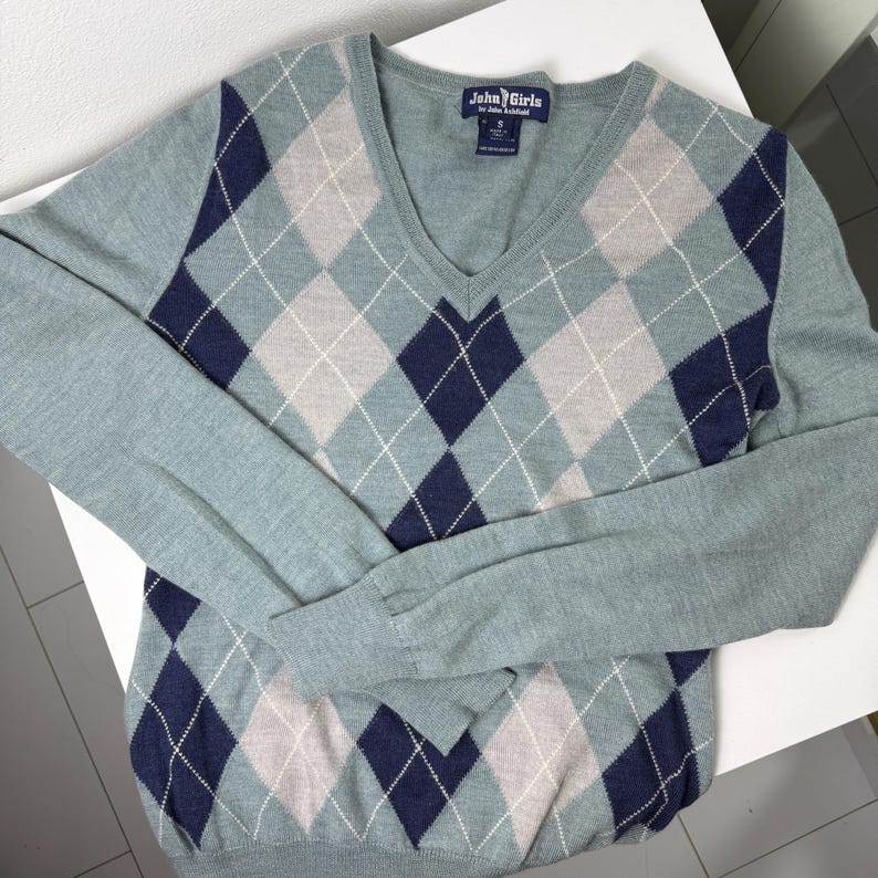 May include: A pale blue V-neck sweater featuring a navy, grey, and white argyle design. The long-sleeved jumper has a label that says "John Girls".