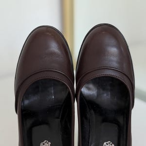 Op de afbeelding: Een paar bruine leren loafers. De schoenen hebben een ronde neus en een lage hak. De zolen zijn zwart en de binnenkant heeft een logo. De schoenen zijn in goede staat en lijken goed onderhouden.