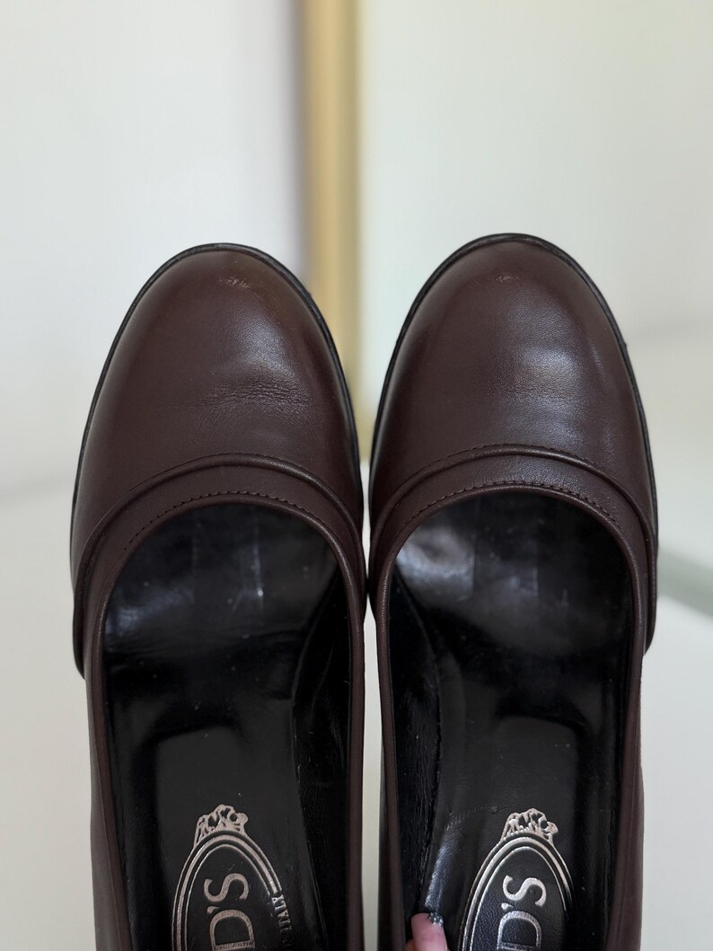 Op de afbeelding: Een paar bruine leren loafers. De schoenen hebben een ronde neus en een lage hak. De zolen zijn zwart en de binnenkant heeft een logo. De schoenen zijn in goede staat en lijken goed onderhouden.