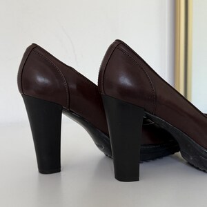 Op de afbeelding: Een paar donkerbruine leren pumps met zwarte blokhakken. De schoenen hebben een klassiek design met een ronde neus en een subtiel naaddetail. De hakken zijn ongeveer 7.6 cm hoog.