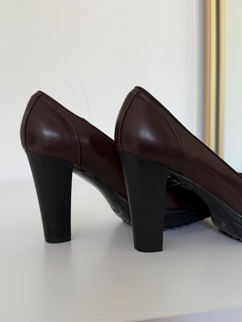 Op de afbeelding: Een paar donkerbruine leren pumps met zwarte blokhakken. De schoenen hebben een klassiek design met een ronde neus en een subtiel naaddetail. De hakken zijn ongeveer 7.6 cm hoog.