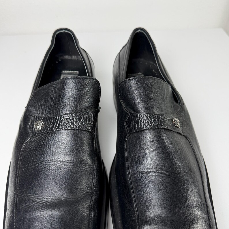 May include: A pair of black leather slip-on shoes. Each shoe features a textured strap across the top with a silver-colored emblem. The shoes have a rounded toe and a low heel, suitable for formal or casual wear.