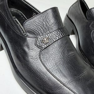 May include: Close-up of a pair of black leather loafers. The shoes have a textured surface and a decorative strap with a silver emblem. The soles are thick and the shoes appear to be new.