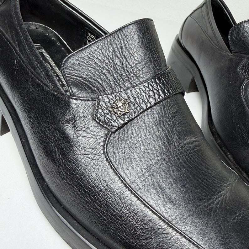 May include: Close-up of a pair of black leather loafers. The shoes have a textured surface and a decorative strap with a silver emblem. The soles are thick and the shoes appear to be new.