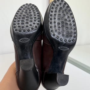 Op de afbeelding: Paar bruine leren laarzen met zwarte zolen. De zolen hebben een textuurontwerp met de merknaam "TOD'S" in reli&euml;f. De hakken zijn effen zwart. De laarzen worden van de zool naar boven getoond.