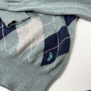 May include: A light blue argyle sweater with navy blue and grey diamond patterns. The sweater has a teal embroidered design near the bottom and a ribbed cuff. The sweater is made of a soft knit material.