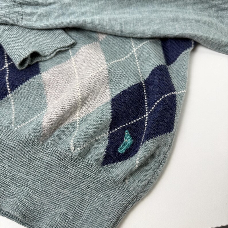 May include: A light blue argyle sweater with navy blue and grey diamond patterns. The sweater has a teal embroidered design near the bottom and a ribbed cuff. The sweater is made of a soft knit material.