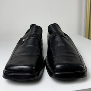 May include: A pair of black leather slip-on shoes. The shoes have a square toe design and a strap across the top with a decorative silver-colored emblem. The soles are thick and black.