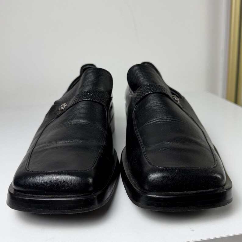 May include: A pair of black leather slip-on shoes. The shoes have a square toe design and a strap across the top with a decorative silver-colored emblem. The soles are thick and black.