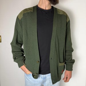May include: An olive green cardigan with suede shoulder patches and two buttons. The cardigan has a ribbed texture and a small pocket with a red embroidered detail. The sleeves have ribbed cuffs.