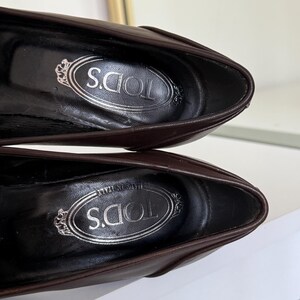 Op de afbeelding: Close-up van twee bruine leren loafers. De binnenkant van elke schoen toont de merknaam "TOD'S" in zilveren letters. De schoenen zijn zo gepositioneerd dat de zolen naar boven wijzen, met het logo van het merk.