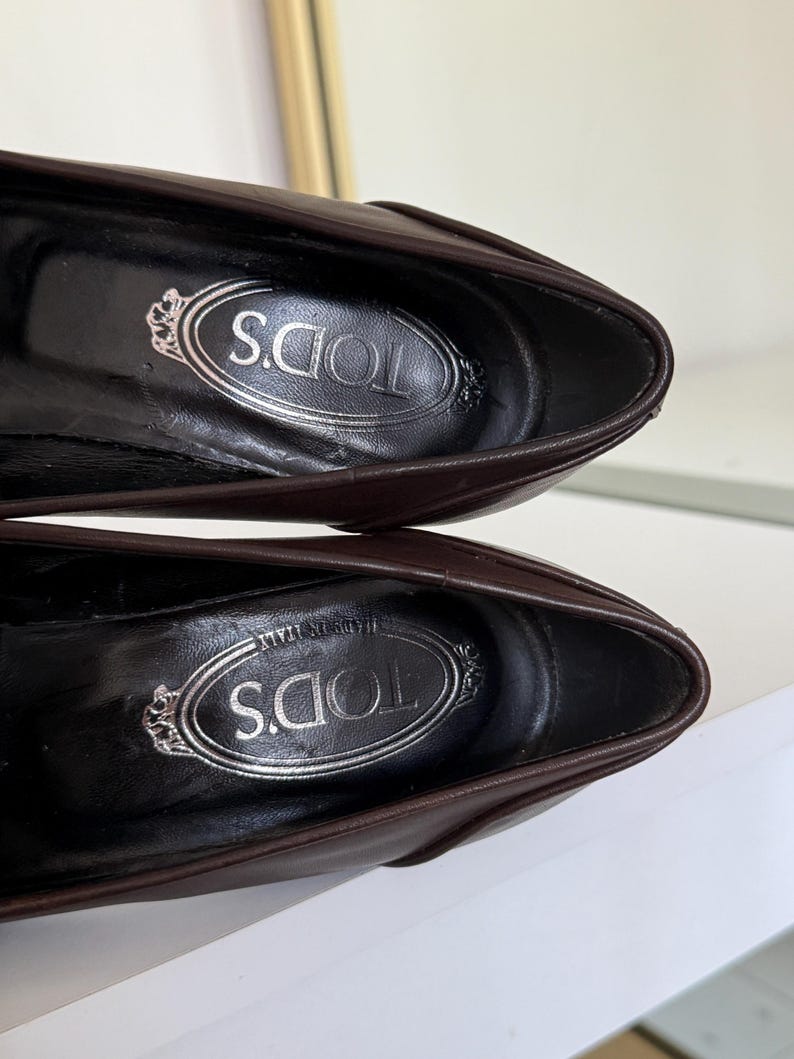 Op de afbeelding: Close-up van twee bruine leren loafers. De binnenkant van elke schoen toont de merknaam "TOD'S" in zilveren letters. De schoenen zijn zo gepositioneerd dat de zolen naar boven wijzen, met het logo van het merk.