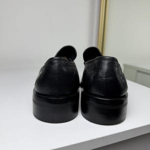 May include: A pair of black leather loafers with a rounded toe and a thick sole. The shoes are displayed from the back, showcasing the smooth texture of the leather and the solid construction of the sole.