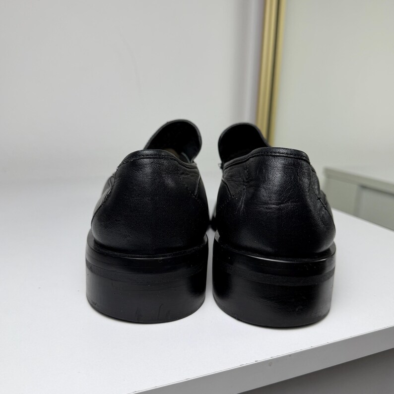 May include: A pair of black leather loafers with a rounded toe and a thick sole. The shoes are displayed from the back, showcasing the smooth texture of the leather and the solid construction of the sole.