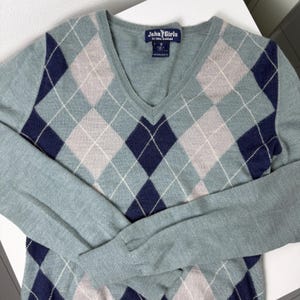 May include: A pale blue V-neck sweater featuring an argyle design in navy, light grey, and white. The long-sleeved sweater has a label that reads "John Girls".