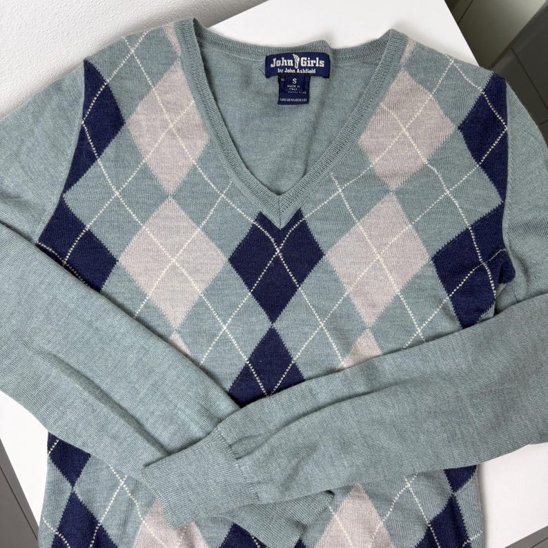 May include: A pale blue V-neck sweater featuring an argyle design in navy, light grey, and white. The long-sleeved sweater has a label that reads "John Girls".