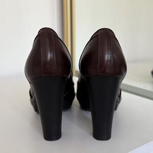 Op de afbeelding: Een paar bruine leren pumps voor dames. De schoenen hebben een donkerbruin bovenwerk en een zwarte blokhak. De hakken zijn ongeveer 10 cm hoog. De schoenen worden op een wit oppervlak gepresenteerd.
