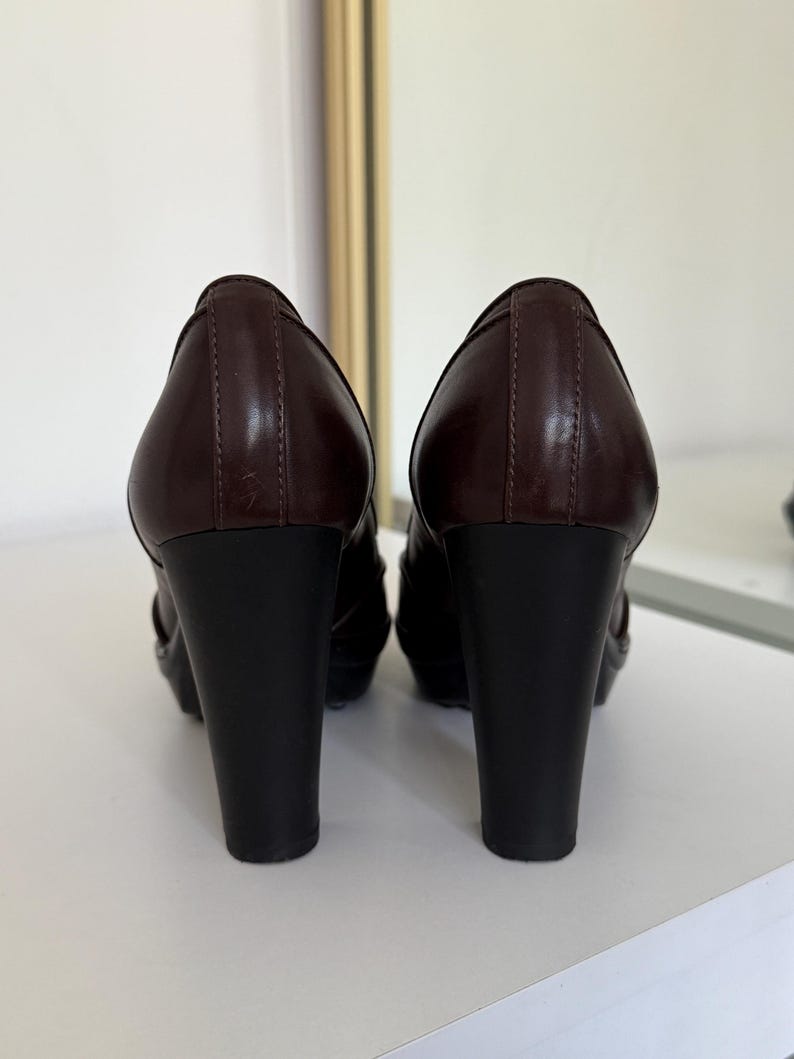 Op de afbeelding: Een paar bruine leren pumps voor dames. De schoenen hebben een donkerbruin bovenwerk en een zwarte blokhak. De hakken zijn ongeveer 10 cm hoog. De schoenen worden op een wit oppervlak gepresenteerd.