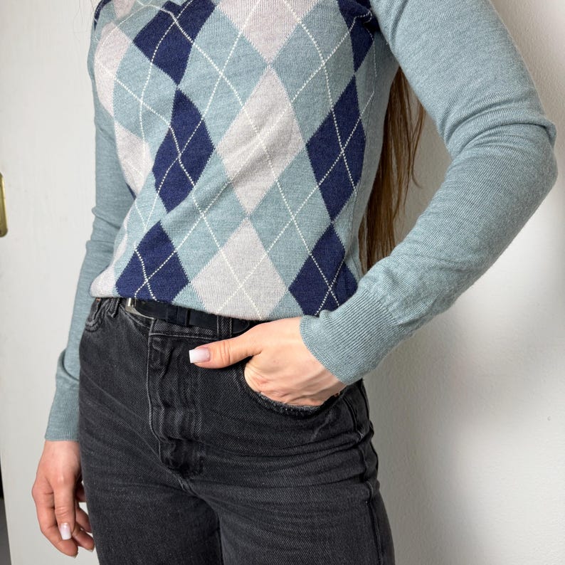 May include: A pale blue sweater featuring an argyle design in navy, grey, and white. The long-sleeved top is worn with black denim jeans.
