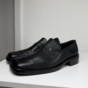 May include: A pair of black leather loafers with a square toe and small heel. The shoes have a strap across the top with a decorative metal emblem. The loafers are sitting on a white surface.