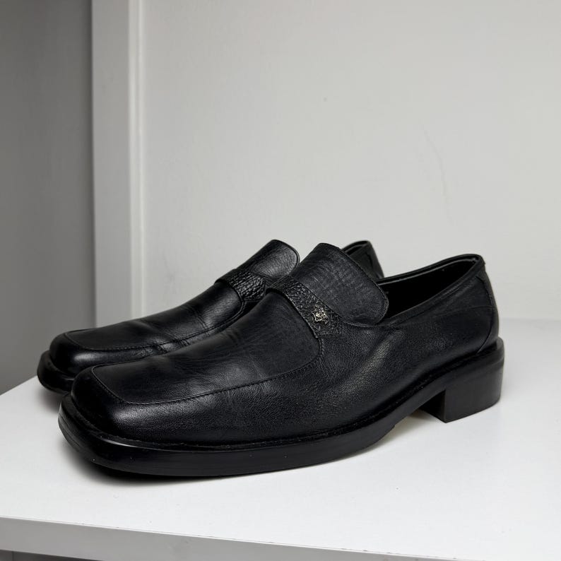 May include: A pair of black leather loafers with a square toe and small heel. The shoes have a strap across the top with a decorative metal emblem. The loafers are sitting on a white surface.