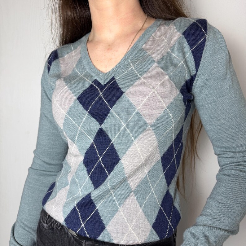 May include: A V-neck sweater with an argyle pattern in shades of blue, navy, grey and white. The long sleeves and the rest of the sweater are a solid blue colour. A classic, timeless design.