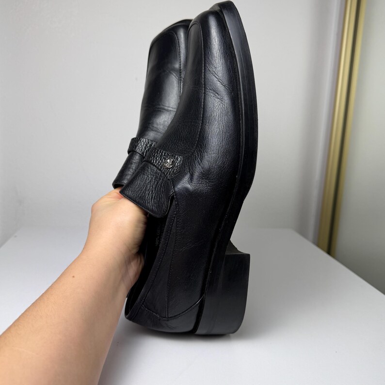 May include: Black leather loafers with a square toe and a small decorative buckle. The shoes have a low, block heel. The loafers are held up by a hand.