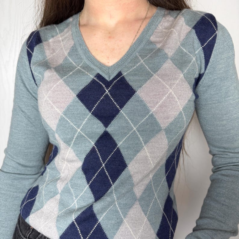 May include: A light blue V-neck sweater featuring an argyle design in blue, grey, and white. The long sleeves are a solid light blue. The image is a close-up of the sweater.