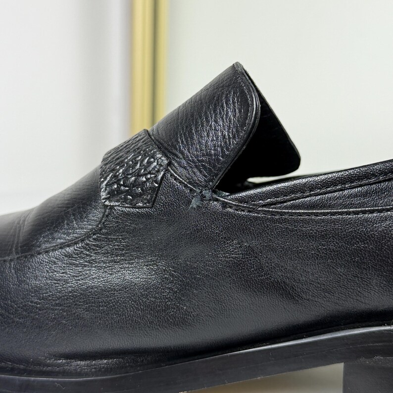 May include: Close-up of a black leather loafer. The shoe features a textured strap across the top and a low heel. The leather has a subtle grain, and the stitching is visible. The overall design is classic and minimalist.