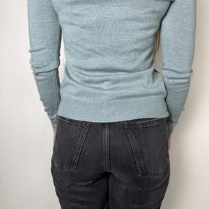 May include: A pale blue sweater with long sleeves and a crew neck is worn with black denim jeans. The jeans have a classic five-pocket design. The person is standing in front of a white wall.