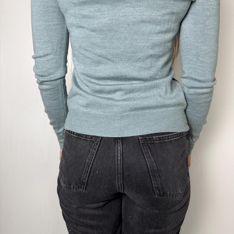 May include: A pale blue sweater with long sleeves and a crew neck is worn with black denim jeans. The jeans have a classic five-pocket design. The person is standing in front of a white wall.