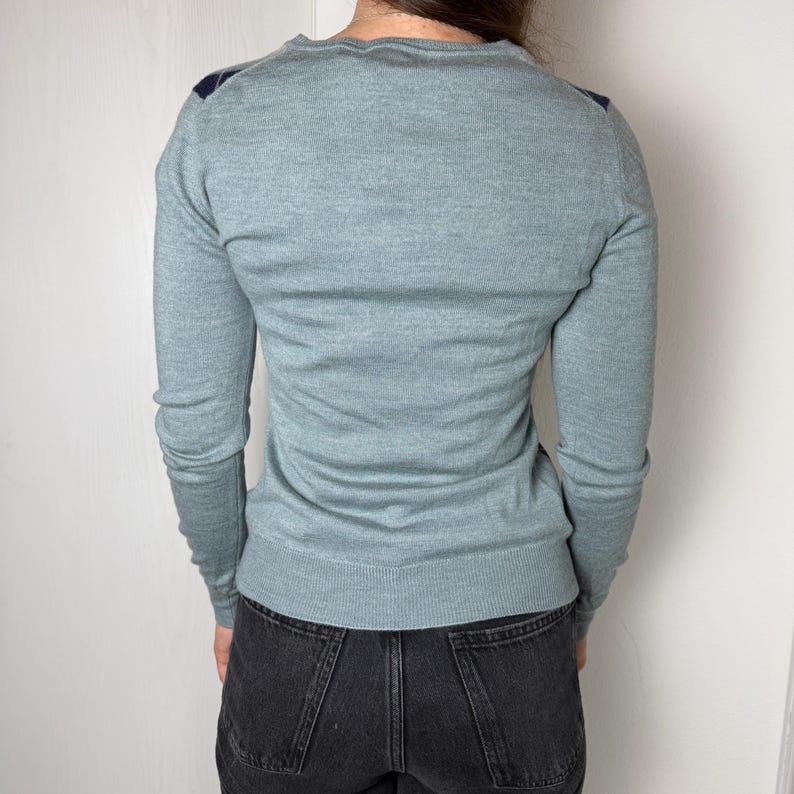May include: A pale blue sweater with long sleeves and a crew neck. Dark blue accents are on the shoulders. The sweater is made of a soft, knitted material. The person is wearing black denim jeans.