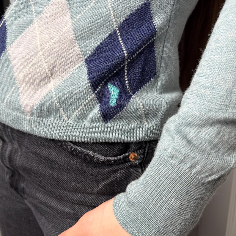 May include: A light blue argyle sweater with a teal logo near the hem. The sweater features a pattern of light grey and navy blue diamonds. The sleeve and hem are ribbed. The sweater is paired with black denim jeans.