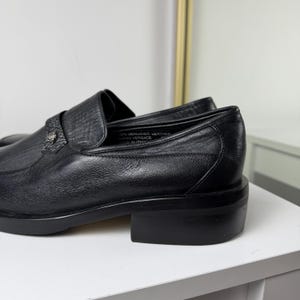 May include: Black leather loafers with a low block heel. The shoes have a textured vamp and a decorative metal emblem. The interior of the shoe is visible, with text that reads "100% Genuine Leather".