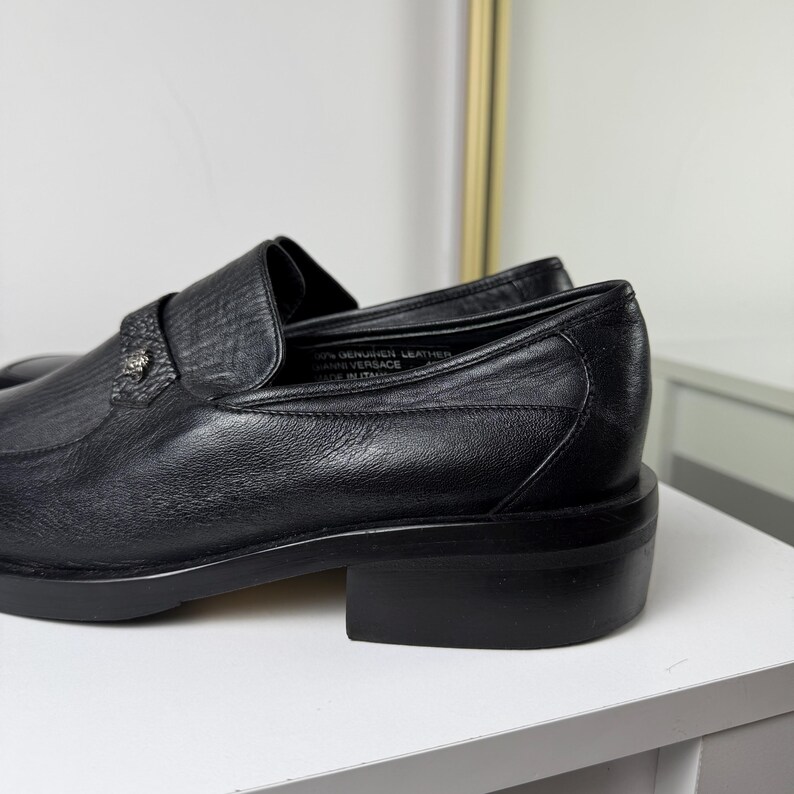 May include: Black leather loafers with a low block heel. The shoes have a textured vamp and a decorative metal emblem. The interior of the shoe is visible, with text that reads "100% Genuine Leather".