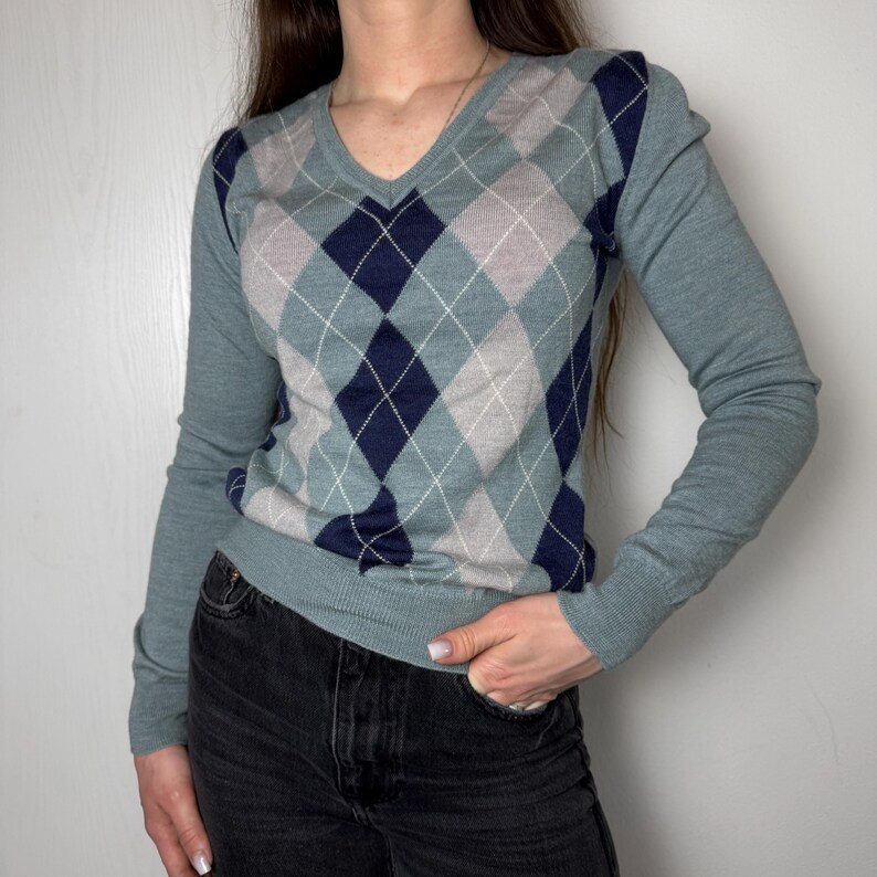 May include: A teal V-neck sweater with long sleeves, featuring a navy, grey, and white argyle pattern. The sweater has a ribbed hem and cuffs. The argyle pattern is made of diamond shapes.