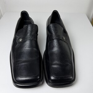 May include: A pair of black leather loafers with square toes. Each shoe features a textured strap across the top with a silver-colored emblem. The shoes are displayed on a white surface.