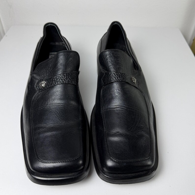May include: A pair of black leather loafers with square toes. Each shoe features a textured strap across the top with a silver-colored emblem. The shoes are displayed on a white surface.