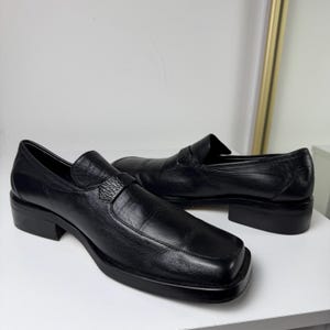 May include: A pair of black leather loafers with a square toe and a low block heel. The shoes have a textured strap across the top and a smooth, polished finish.