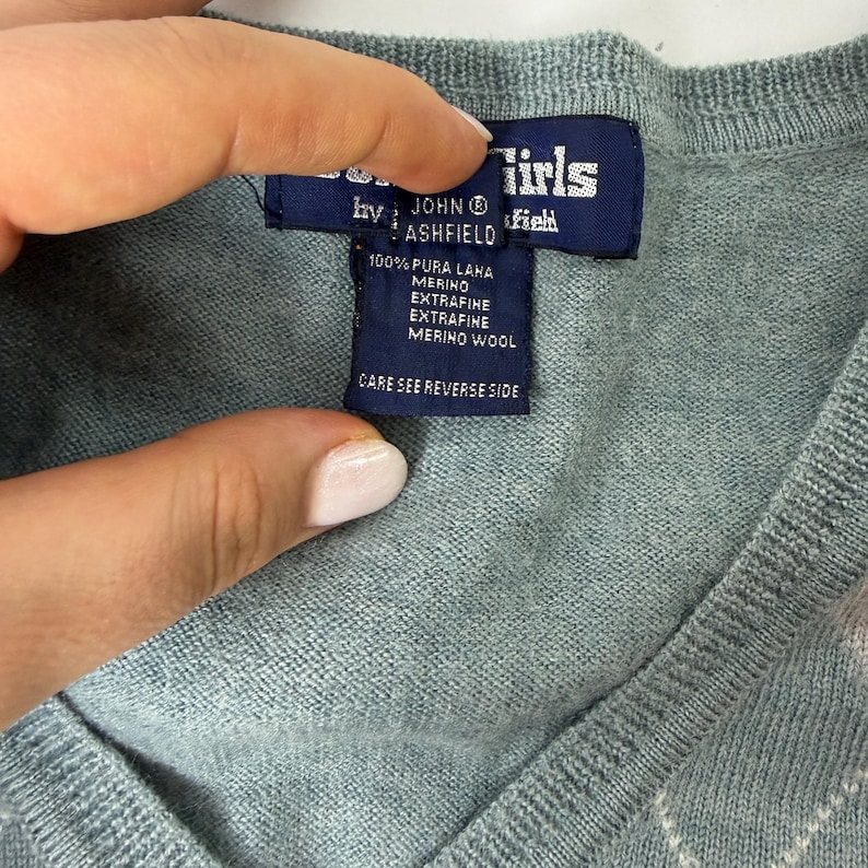 May include: A close-up of a pale blue sweater with a V-neck. A navy blue label shows the brand "John Ashfield" and the text "100% Pura Lana Merino Extrafine Merino Wool". The label also includes the text "Care see reverse side".