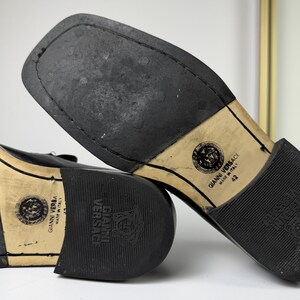 May include: Black leather dress shoes with square toes. The soles are black rubber with the "Gianni Versace Made in Italy" logo and size 42. The shoes are in a close-up shot, showcasing the soles.