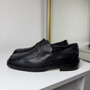 May include: Black leather loafers with a square toe and a low heel. The shoes have a textured vamp and a decorative strap across the top. The shoes are displayed on a white surface.