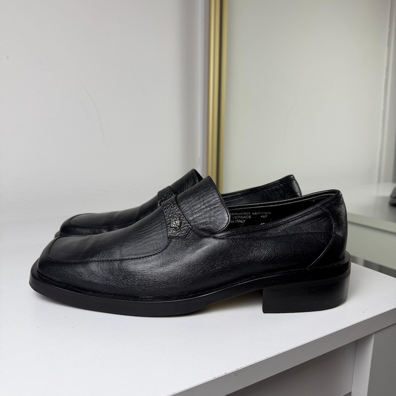 May include: Black leather loafers with a square toe and a low heel. The shoes have a textured vamp and a decorative strap across the top. The shoes are displayed on a white surface.
