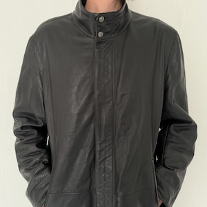 May include: A black leather jacket with a high collar and two button fastening. The jacket has a textured finish and two front pockets. The sleeves are long, and the jacket appears to be a mid-length coat.