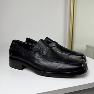 May include: A pair of black leather slip-on shoes with a square toe and a small block heel. The shoes have a decorative strap across the top with a silver-colored emblem. The shoes are displayed on a white surface.